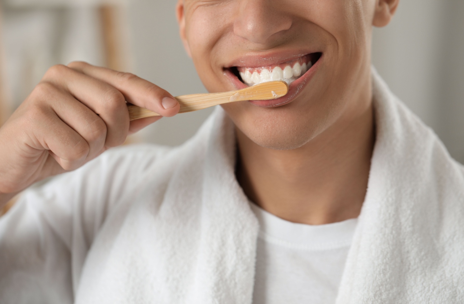 A man smiles brightly while brushing his teeth in front of a bathroom mirror.