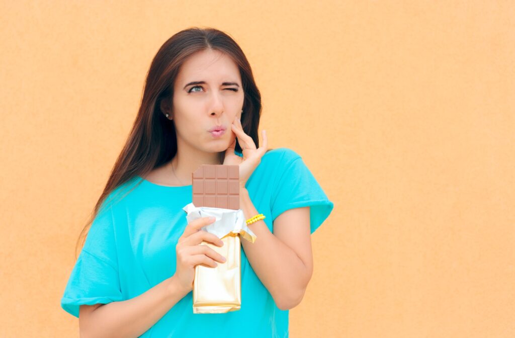 A girl holding a chocolate bar with one hand and grabbing her cheek with the other due to tooth sensitivity when eating sugar