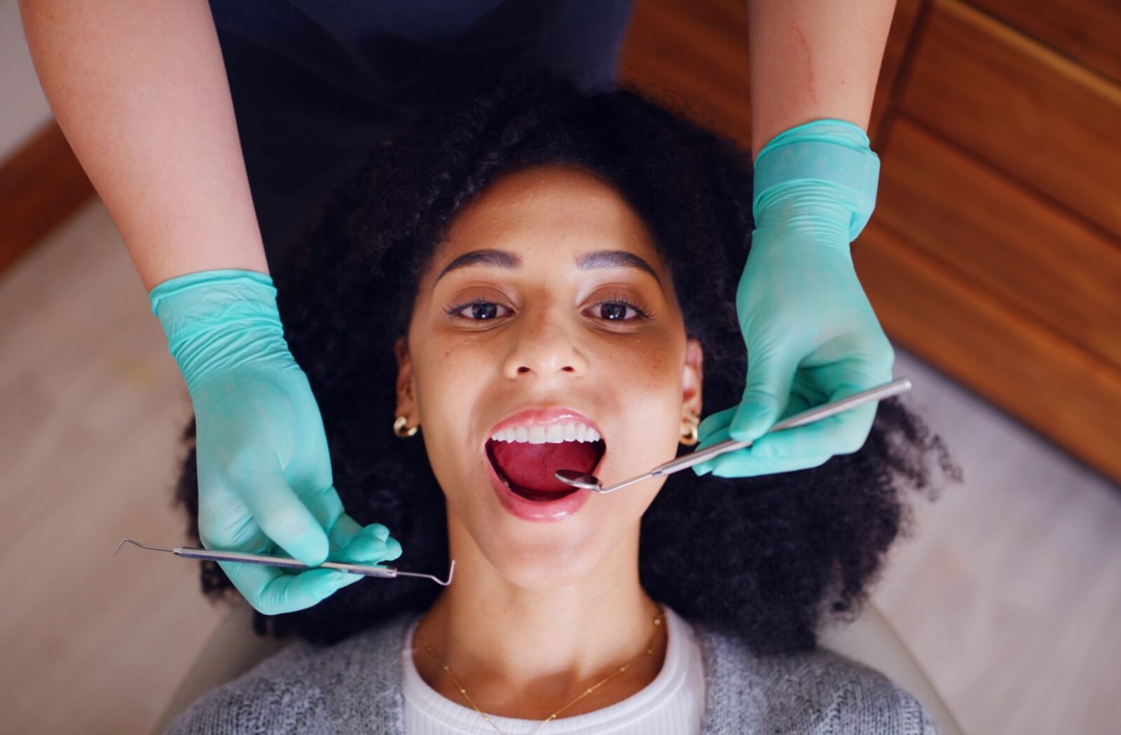 A top-down view of a dental patient with their mouth open while a provider in teal gloves performs an exam using dental tools.